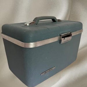 May include: A rectangular, slate blue hardshell travel case with a textured finish. It features a top handle, a silver-toned metal trim around the center, and a central locking latch. The word Starlite is printed on a small silver plate on the front.