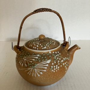May include: A round, textured tan ceramic teapot featuring a raised white and light green floral pattern on the body and lid. The teapot includes a curved spout and a brown woven rattan handle attached to two metal loops on the sides.