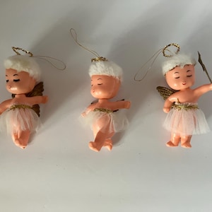 May include: Three vintage angel ornaments. Each has a peach-colored body, white fluffy hair, and gold halo. They wear a white tulle skirt with gold trim and have gold wings. One angel holds a small gold wand.