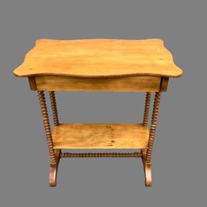May include: A light brown, antique side table with a scalloped top and a lower shelf. The table features turned legs and a drawer. The table is made of wood and has a warm, natural finish. The table is approximately 30 inches tall.