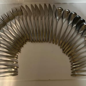 May include: Arrangement of stainless steel cutlery, including forks, knives, and spoons, forming an arc shape. The silverware is displayed on a white surface, showcasing a complete set of dining utensils.