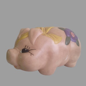 May include: A pink ceramic piggy bank with a yellow bow and a purple flower on its side.