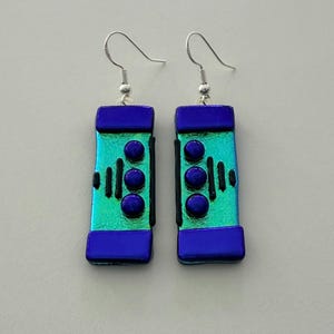 May include: A pair of handmade dichroic glass earrings. Each earring features a rectangular shape with a teal and blue color scheme. The earrings have a silver hook and are adorned with three blue circular accents and black vertical lines.