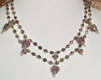 Peut inclure: Un collier à deux rangs avec des perles iridescentes violettes et argentées. Le collier présente plusieurs perles en forme de cœur pendantes avec des accents de fil d'or.