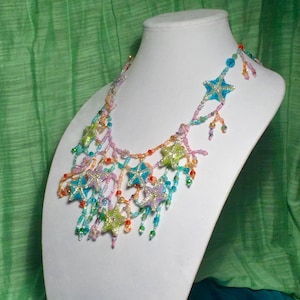 May include: A colourful beaded necklace with a cascading design featuring multiple star-shaped beads in shades of blue, green, pink, and orange. The necklace is displayed on a white mannequin bust.