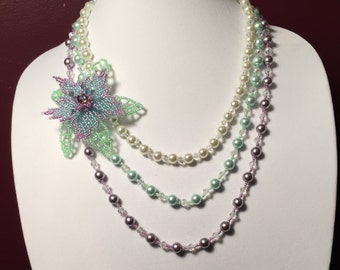 Peut inclure: Un collier multi-rangs avec un pendentif en forme de fleur délicate. Le collier présente trois rangs de perles dans des tons de blanc, vert et violet.