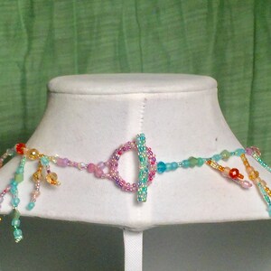 Peut inclure: Un collier de perles multicolores avec un fermoir &agrave; glissi&egrave;re. Le collier pr&eacute;sente une vari&eacute;t&eacute; de perles dans des tons de rose, bleu, vert et orange. Les perles sont dispos&eacute;es en cascade, cr&eacute;ant un look fantaisiste et &eacute;l&eacute;gant.