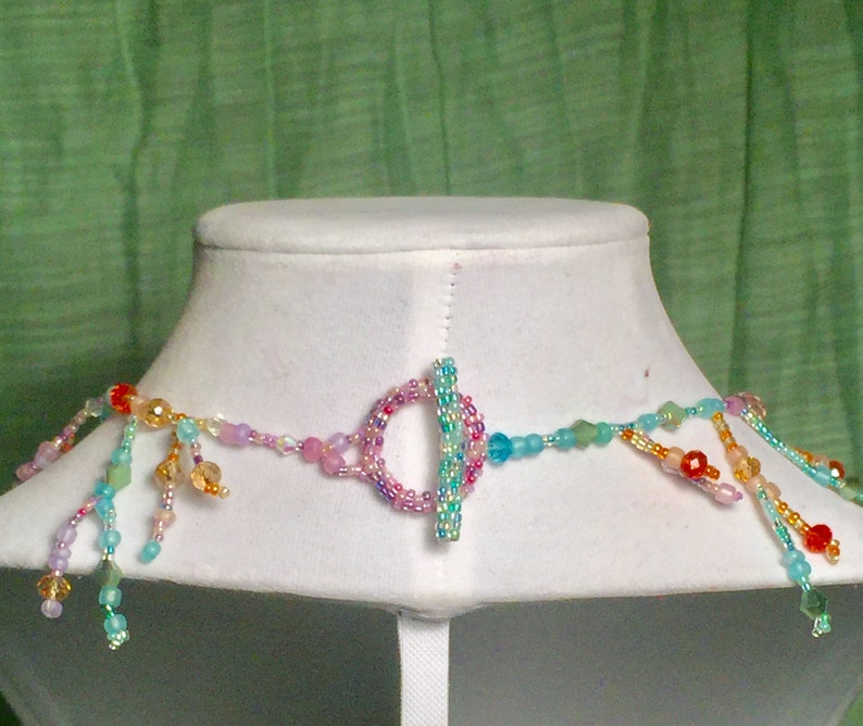 Peut inclure: Un collier de perles multicolores avec un fermoir &agrave; glissi&egrave;re. Le collier pr&eacute;sente une vari&eacute;t&eacute; de perles dans des tons de rose, bleu, vert et orange. Les perles sont dispos&eacute;es en cascade, cr&eacute;ant un look fantaisiste et &eacute;l&eacute;gant.