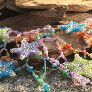 Peut inclure: Un collier de perles color&eacute; avec des breloques en forme d'&eacute;toile. Le collier est compos&eacute; de perles de diff&eacute;rentes nuances de bleu, vert, rose et violet. Les breloques en forme d'&eacute;toile sont argent&eacute;es avec un centre bleu clair, vert ou violet.