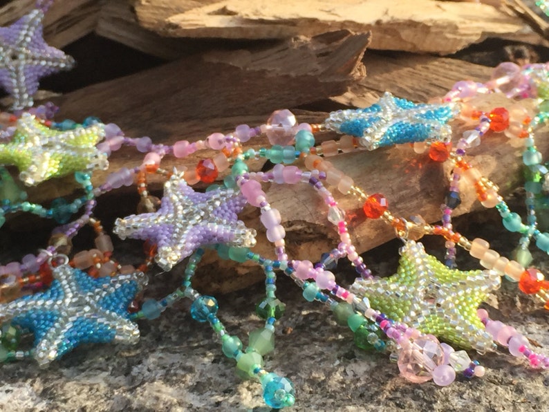 Peut inclure: Un collier de perles color&eacute; avec des breloques en forme d'&eacute;toile. Le collier est compos&eacute; de perles de diff&eacute;rentes nuances de bleu, vert, rose et violet. Les breloques en forme d'&eacute;toile sont argent&eacute;es avec un centre bleu clair, vert ou violet.