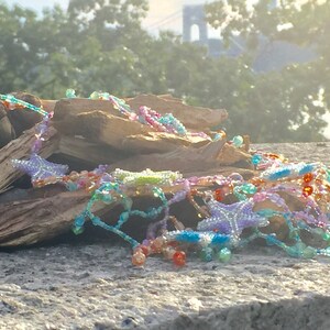 Peut inclure: Un collier de perles color&eacute; avec des breloques en forme d'&eacute;toile, reposant sur une pile de bois us&eacute;. Le collier pr&eacute;sente une vari&eacute;t&eacute; de couleurs, notamment le rose, le bleu, le vert et l'orange.