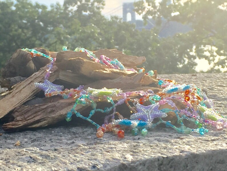 Peut inclure: Un collier de perles color&eacute; avec des breloques en forme d'&eacute;toile, reposant sur une pile de bois us&eacute;. Le collier pr&eacute;sente une vari&eacute;t&eacute; de couleurs, notamment le rose, le bleu, le vert et l'orange.