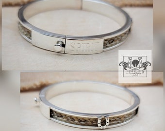 May include: A silver-toned hinged bangle bracelet with a braided horsehair inlay. The word "SPIRIT" is engraved on the clasp. A small horseshoe charm is visible. The bracelet is displayed on a light-colored surface.