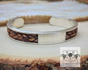 Könnte beinhalten: Ein silbernes Armreif-Armband mit geflochtener Pferdehaareinlage. Das Armband trägt den eingravierten Namen "VINCENT" auf einer rechteckigen Platte. Das Pferdehaar ist eine Mischung aus braunen, beigen und schwarzen Farben. Das Armband wird auf einer Holzoberfläche präsentiert.