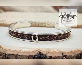 Può includere: Bracciale a polsino in argento con una fascia di crine di cavallo intrecciato. Un piccolo ciondolo a forma di ferro di cavallo in argento è centrato sul davanti. Il bracciale è esposto su una superficie di legno. Il logo "Horse Hair Creations by Dale" è sullo sfondo.