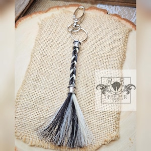 May include: A keychain featuring a braided horse hair design in black and white. The keychain has a silver-colored clasp and ring. The horse hair is braided into a long, tassel-like shape. The background is a burlap-like material.