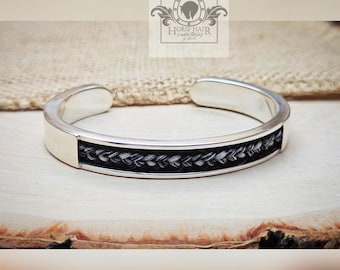 May include: A silver cuff bracelet with a braided horse hair inlay. The horse hair is black and white, woven in a detailed pattern. The bracelet has a rectangular design and is displayed on a wooden surface.