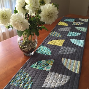 May include: A grey table runner with a geometric pattern of teal, yellow and white half-circles. The runner is quilted and has a subtle texture.
