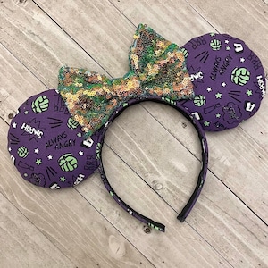 May include: Purple and green mouse ears with a green and gold sequin bow. The ears have a pattern of green fists, stars, and the words "SMASH", "ALWAYS ANGRY", and "Hulk".