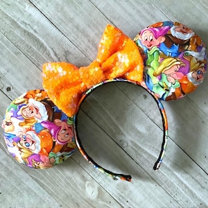 May include: Orange sequin bow headband with a fabric design of the seven dwarfs from Snow White. The headband is made of black felt.