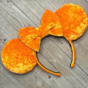 May include: A pair of orange velvet Minnie Mouse ears with a large bow. The ears are made of a soft, plush fabric and are perfect for any Disney fan.