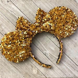 Op de afbeelding: Gouden Minnie Mouse oren met pailletten en een fluwelen strik. De oren zijn bedekt met gouden pailletten en de strik is gemaakt van zacht, pluche fluweel.