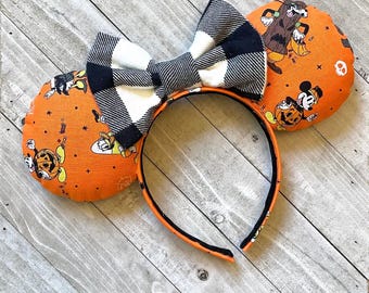 May include: Orange and black Halloween-themed Minnie Mouse ears with a black and white plaid bow. The ears feature a variety of Halloween characters, including Mickey Mouse, Minnie Mouse, and Donald Duck.