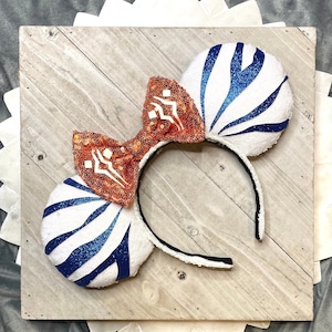 以下が含まれることがあります： 白と青のストライプのミッキーマウスイヤーヘッドバンドと、オレンジ色のスパンコールリボン。耳には青いグリッターのデザイン。ヘッドバンドは黒で、白いトリムが付いています。リボンには白いデザインがあります。