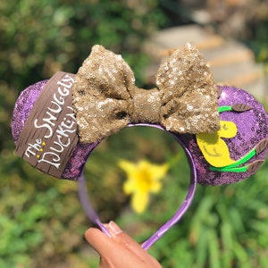 May include: Purple Minnie Mouse ears with a gold sequin bow and a brown sign that says "The Snuggly Duckling".