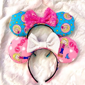 May include: A pair of pink and blue mouse ears with a cartoon character on each ear. The ears are decorated with stars, hearts, and the character's name. The headband is black and has a white bow.