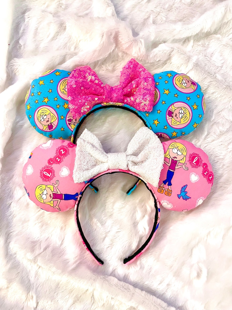 May include: A pair of pink and blue mouse ears with a cartoon character on each ear. The ears are decorated with stars, hearts, and the character's name. The headband is black and has a white bow.