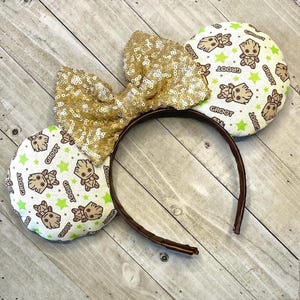 May include: A pair of brown headband mouse ears with a gold sequined bow. The ears are covered in a white fabric with a repeating pattern of green stars and brown cartoon characters with the word "Groot" written on them.