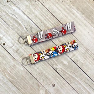 Puede incluir: Dos llaveros con correas de tela. Uno presenta un diseño de dibujos animados de Spiderman con el texto "Soy solo un amigable vecino Spider-Man" y "entrenamiento de lealtad". El otro presenta un diseño de dibujos animados de varios personajes de Marvel.