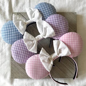May include: Three pairs of gingham mouse ears with white lace bows. The ears are in blue, purple, and pink gingham.