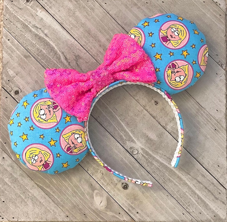 May include: Pink sequined bow headband with blue fabric ears featuring a cartoon character with yellow hair and blue eyes. The ears are covered in yellow stars.