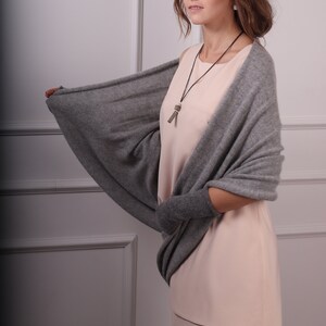 May include: A woman wearing a light pink dress with a gray cashmere wrap. The wrap has a long, flowing design and is draped over her shoulders. She is wearing a silver necklace with a small pendant.