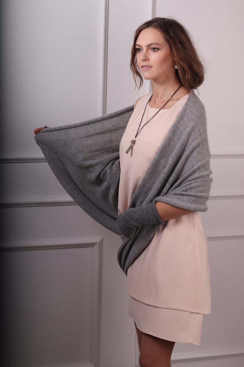 May include: A woman wearing a light pink dress with a gray cashmere wrap. The wrap has a long, flowing design and is draped over her shoulders. She is wearing a silver necklace with a small pendant.