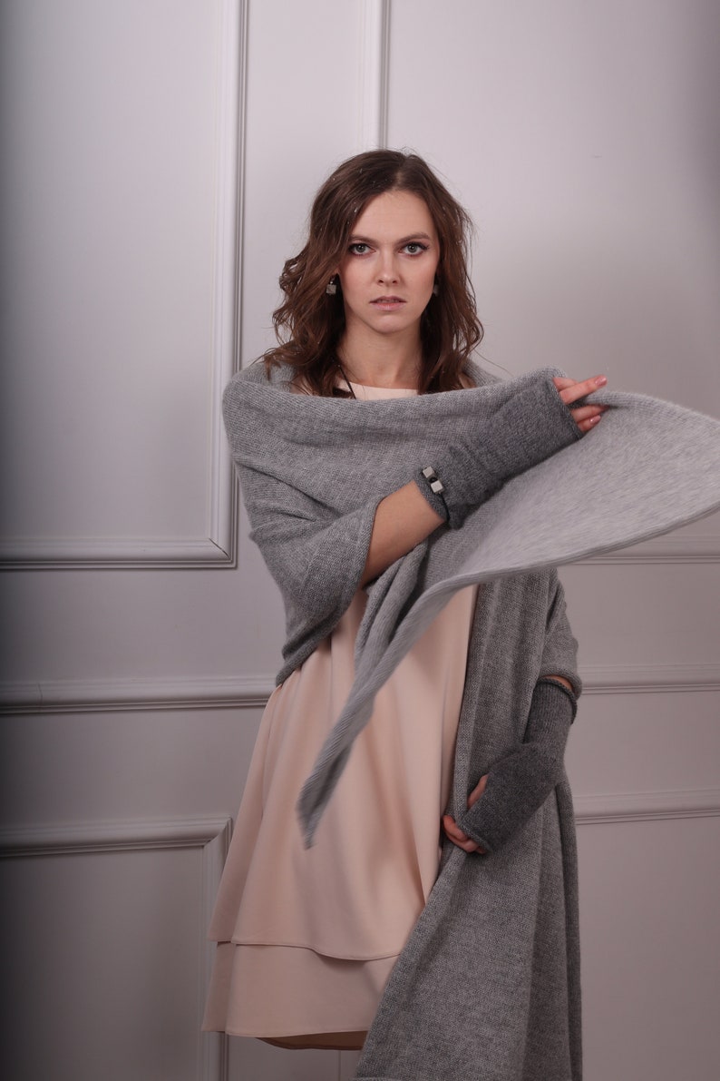May include: A woman wearing a gray cashmere scarf and matching fingerless gloves. The scarf is draped around her shoulders and the gloves are on her hands. She is wearing a light pink dress.