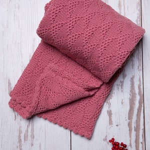 Pode incluir: Uma manta de crochê rosa com uma borda festonada está dobrada sobre uma superfície de madeira branca. Um ramo de bagas vermelhas está no canto inferior direito da imagem.