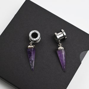 May include: Two silver ear gauges with amethyst stone plugs. The gauges are on a black background.