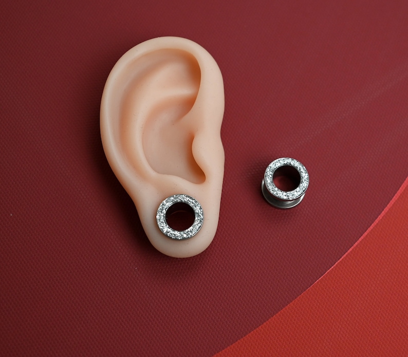 Puede incluir: Un par de expansores de oreja plateados con un anillo de piedras claras y brillantes. Uno est&aacute; insertado en un modelo de oreja color carne, y el otro est&aacute; colocado por separado sobre un fondo rojo.