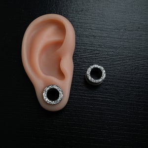 Puede incluir: Un par de expansores de oreja plateados con un anillo de piedras claras y brillantes en el borde exterior. Uno est&aacute; insertado en una oreja prot&eacute;sica, el otro est&aacute; colocado sobre una superficie negra.