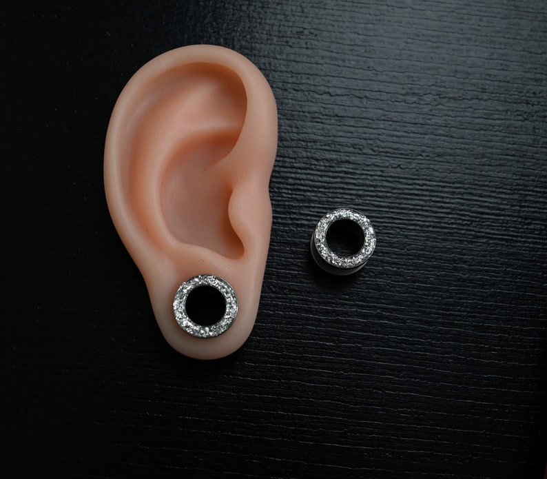 Puede incluir: Un par de expansores de oreja plateados con un anillo de piedras claras y brillantes en el borde exterior. Uno est&aacute; insertado en una oreja prot&eacute;sica, el otro est&aacute; colocado sobre una superficie negra.
