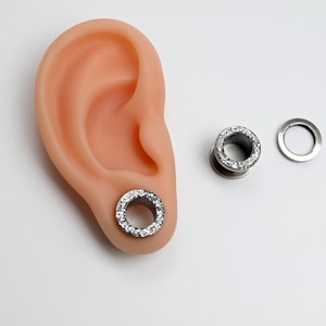 Puede incluir: Un par de expansores de oreja plateados con un anillo de piedras transparentes y brillantes en el borde exterior. Uno est&aacute; insertado en una oreja prot&eacute;sica, el otro est&aacute; separado, junto con una pieza trasera a juego. Fondo blanco.