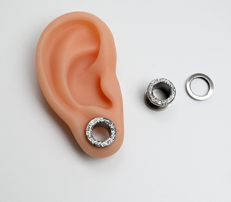 Puede incluir: Un par de expansores de oreja plateados con un anillo de piedras transparentes y brillantes en el borde exterior. Uno est&aacute; insertado en una oreja prot&eacute;sica, el otro est&aacute; separado, junto con una pieza trasera a juego. Fondo blanco.