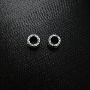 Puede incluir: Dos expansores de oreja plateados con dise&ntilde;o circular. Cada uno est&aacute; adornado con peque&ntilde;as piedras transparentes y brillantes en el borde exterior. Se presentan sobre un fondo oscuro y texturizado.