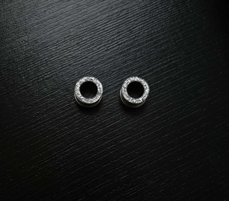 Puede incluir: Dos expansores de oreja plateados con dise&ntilde;o circular. Cada uno est&aacute; adornado con peque&ntilde;as piedras transparentes y brillantes en el borde exterior. Se presentan sobre un fondo oscuro y texturizado.