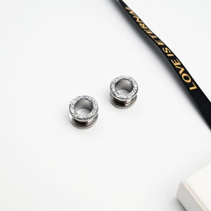 Puede incluir: Dos expansores de oreja plateados, cada uno con un anillo de piedras claras y brillantes en el borde. Los expansores son circulares con un centro hueco. El fondo es blanco, con una cinta negra con el texto dorado "LOVE IS ETERNAL".