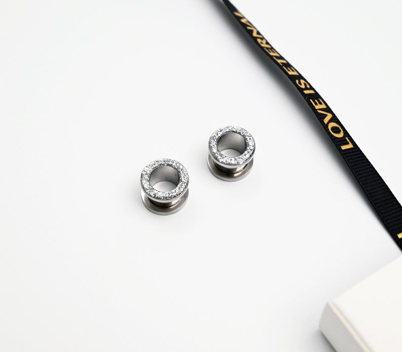 Puede incluir: Dos expansores de oreja plateados, cada uno con un anillo de piedras claras y brillantes en el borde. Los expansores son circulares con un centro hueco. El fondo es blanco, con una cinta negra con el texto dorado "LOVE IS ETERNAL".