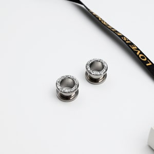 Puede incluir: Dos expansores de oreja plateados con un anillo de peque&ntilde;os cristales transparentes en el borde exterior. Los expansores son circulares con un dise&ntilde;o acampanado. Una cinta negra con texto dorado que dice "LOVE IS ETERNAL" en el fondo.